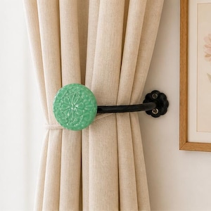 Puede incluir: Un recogedor de cortina decorativo con un acento redondo de cer&aacute;mica verde claro con un dise&ntilde;o floral. El recogedor tiene un brazo de metal negro y un soporte de pared negro en forma de flor. Las cortinas son de color beige claro.
