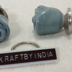 May include: Two light blue ceramic rose-shaped drawer knobs with silver metal bases and screws. The knobs are on a white surface with a brown rectangular tag that says "KRAFTBYINDIA".