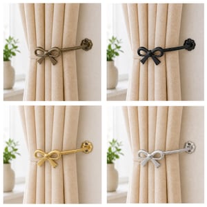 May include: Four curtain tiebacks in different metallic finishes: bronze, black, gold, and silver. Each tieback features a bow-shaped design and a decorative floral end, holding beige curtains. The background includes small white vases with green plants.