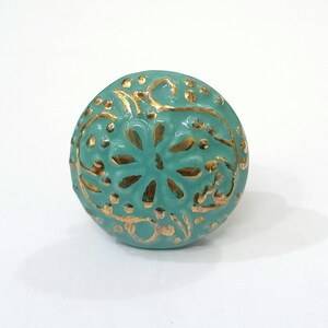 Peut inclure: Un bouton en c&eacute;ramique bleu turquoise avec un motif floral dor&eacute;. Le bouton est rond et pr&eacute;sente un motif en relief.