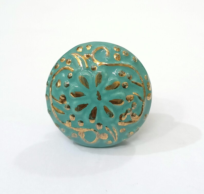 Peut inclure: Un bouton en c&eacute;ramique bleu turquoise avec un motif floral dor&eacute;. Le bouton est rond et pr&eacute;sente un motif en relief.