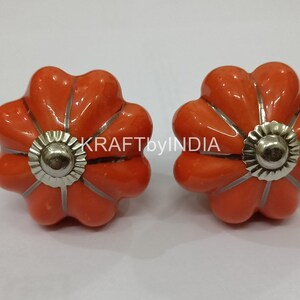 May include: Two orange ceramic flower-shaped cabinet knobs with silver metal accents. KRAFTbyINDIA is printed on the white background.