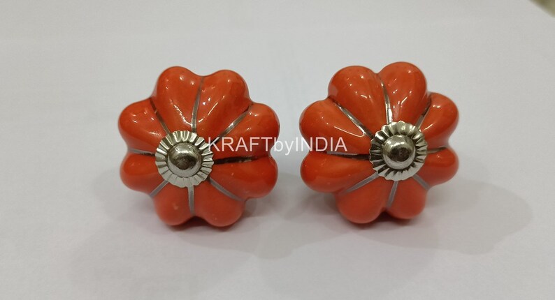 May include: Two orange ceramic flower-shaped cabinet knobs with silver metal accents. KRAFTbyINDIA is printed on the white background.