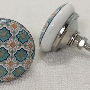 以下が含まれることがあります： 白い背景に青、緑、オレンジの幾何学模様が施されたセラミック製のキャビネットノブ2個。ノブには銀色の金属製のネジが付いています。