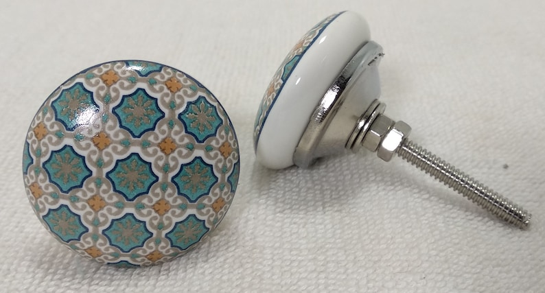 以下が含まれることがあります： 白い背景に青、緑、オレンジの幾何学模様が施されたセラミック製のキャビネットノブ2個。ノブには銀色の金属製のネジが付いています。