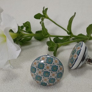 以下が含まれることがあります： 青、緑、オレンジの幾何学模様が施されたセラミック製の引き出しノブが2つ。ノブは丸型で、銀色のネジが付いています。