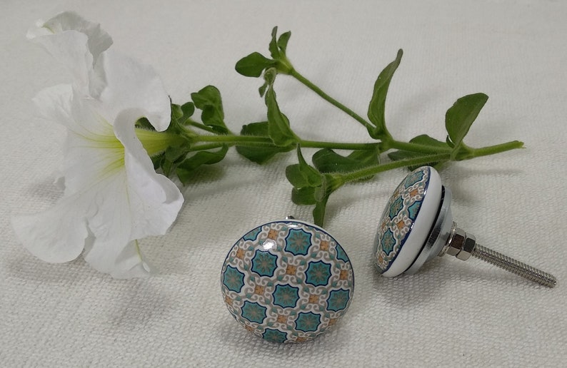 以下が含まれることがあります： 青、緑、オレンジの幾何学模様が施されたセラミック製の引き出しノブが2つ。ノブは丸型で、銀色のネジが付いています。