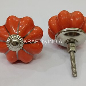 May include: Two orange ceramic pumpkin-shaped cabinet knobs with silver accents and a screw. KRAFTbyINDIA is visible in the background.