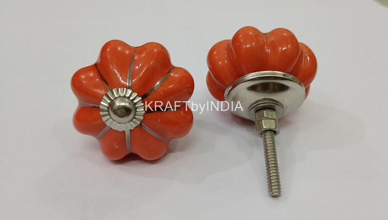 May include: Two orange ceramic pumpkin-shaped cabinet knobs with silver accents and a screw. KRAFTbyINDIA is visible in the background.