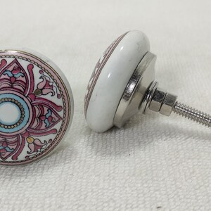 May include: Two white ceramic cabinet knobs with a floral design in pink, blue, and gold. The knobs have a silver metal base and a threaded screw for installation. The design is a circular pattern.