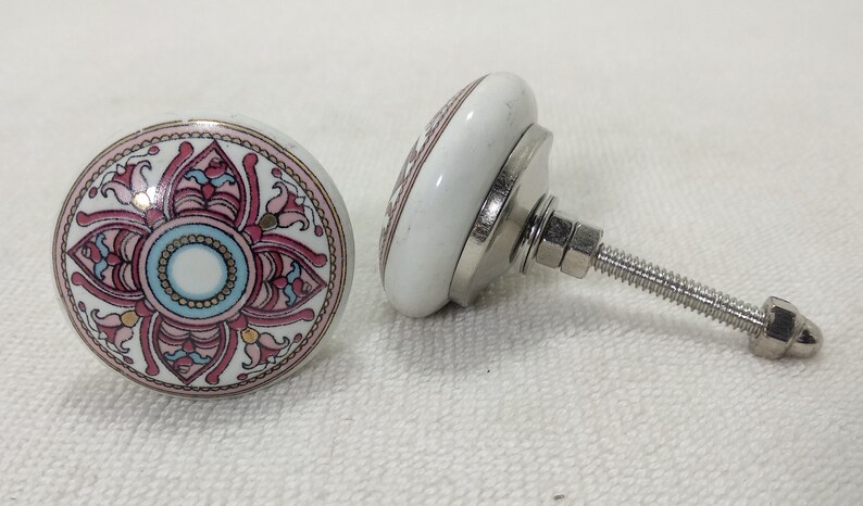 May include: Two white ceramic cabinet knobs with a floral design in pink, blue, and gold. The knobs have a silver metal base and a threaded screw for installation. The design is a circular pattern.