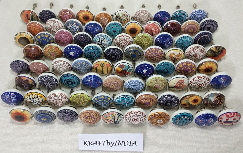 May include: A collection of 48 ceramic cabinet knobs featuring a variety of colourful patterns and designs. The knobs are round and have a silver metal screw. The knobs are arranged in rows on a white surface. The text "KRAFTbyINDIA" is visible at the bottom of the image.