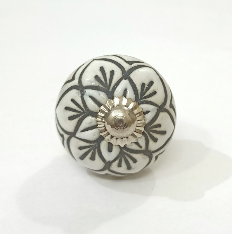 May include: White ceramic knob with a black floral design. The knob has a silver metal center.