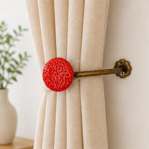 Puede incluir: Un recogedor de cortina redondo rojo con un dise&ntilde;o floral. El recogedor es de cer&aacute;mica y tiene un brazo de metal color bronce que se fija a la pared. La cortina es de color beige claro.