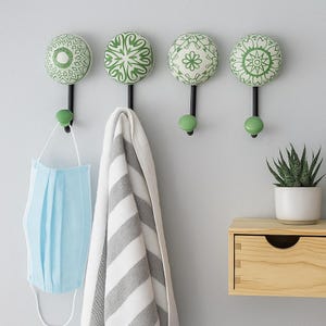 May include: Four wall hooks with decorative green and white ceramic knobs. The hooks are black with a green ball at the bottom. A light blue face mask and a gray and white striped towel hang from the hooks. A small potted plant and a wooden drawer are also visible.