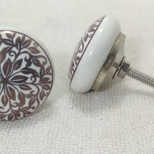 May include: Two white ceramic cabinet knobs with a brown floral design. The knobs have a silver metal base and a screw for attaching to a cabinet door.