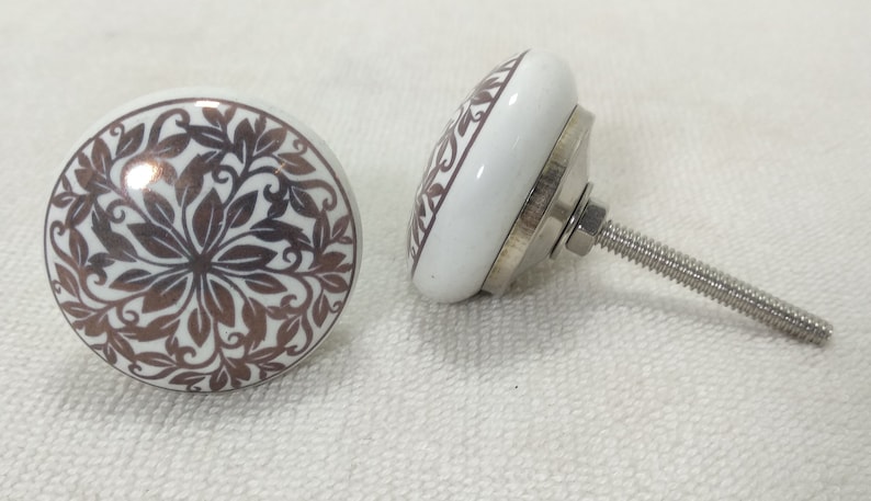 May include: Two white ceramic cabinet knobs with a brown floral design. The knobs have a silver metal base and a screw for attaching to a cabinet door.