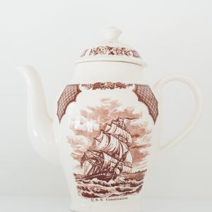 Op de afbeelding: Crèmekleurige keramische theepot met een bruine illustratie van een zeilschip en de tekst "U.S.S. Constitution". De theepot heeft een deksel met bloemendetails en een gebogen handvat. Het ontwerp doet denken aan vintage servies.
