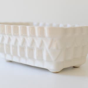 Può includere: Vaso in ceramica bianca con un design geometrico. Il vaso ha una forma rettangolare e un bordo leggermente rialzato.