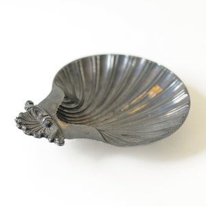 Puede incluir: Un plato de color plateado en forma de concha con un asa decorativa. El plato está hecho de metal y tiene un acabado liso y brillante.