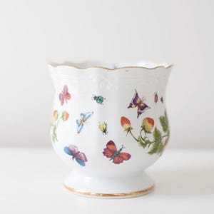 May include: White porcelain vase with a scalloped rim and a gold trim. The vase is decorated with a floral pattern of butterflies, strawberries, and ladybugs.