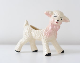 May include: A ceramic lamb planter in off-white with pink accents. The lamb has a textured body, black hooves, and a pink bow. Its face features black eyes and a red mouth. This decorative planter is ideal for small plants or flowers.
