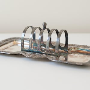 May include: A vintage silver-plated toast rack with a rectangular base and a curved handle. The rack has slots for holding toast upright. The base has a decorative, ornate edge. The piece is tarnished, giving it a vintage look.