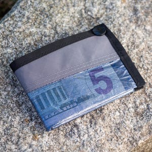 Peut inclure: Un portefeuille bifold gris et noir avec une fermeture à bouton-pression. Le portefeuille a une fenêtre en plastique transparent révélant un billet bleu avec le chiffre 5 imprimé en violet. Le portefeuille est posé sur une surface en pierre grise.