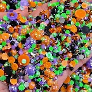 May include: A large assortment of colorful, iridescent, and opaque round rhinestones in various sizes. The rhinestones are in shades of green, orange, purple, black, and white.