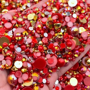 May include: A large assortment of red, gold, and clear rhinestones and beads. The rhinestones are various sizes and shapes, including round, oval, and square. The beads are mostly round and have a pearlescent finish.
