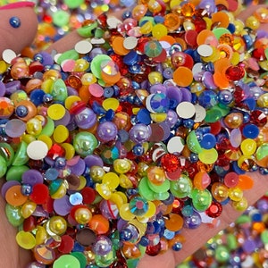 May include: A handful of colorful, assorted craft embellishments. The mix includes various shapes and sizes of rhinestones, pearls, and flat-backed gems in shades of red, orange, yellow, green, blue, and purple. Some have an iridescent sheen.