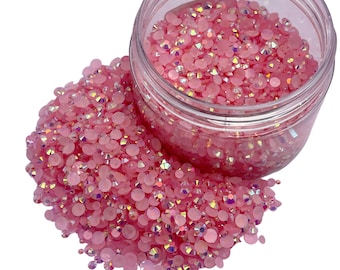 Pode incluir: Um frasco de plástico transparente cheio de strass rosa iridescentes. Os strass estão espalhados sobre uma superfície branca.