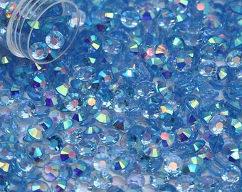 Pode incluir: Uma imagem de close-up de uma grande quantidade de pequenos strass iridescentes azuis claros. Os strass estão espalhados sobre uma superfície branca e parecem estar saindo de um recipiente de plástico transparente.