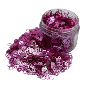 May include: A clear plastic jar filled with pink and silver glitter in the shape of small circles. The glitter is spilling out of the jar onto a white background.