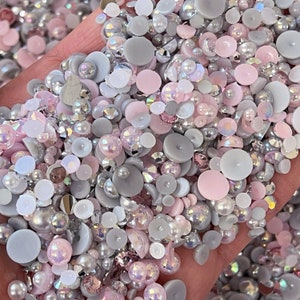 May include: A mix of small, round, and flat decorative embellishments in shades of pink, white, and gray. The embellishments are made of plastic or glass and have a variety of textures.