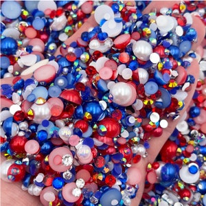 May include: A close-up of a hand holding a variety of red, white, and blue rhinestones and pearls. The rhinestones are in various shapes and sizes, and the pearls are white and round.