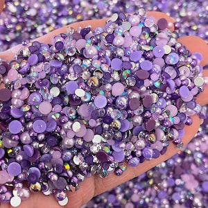 May include: A large assortment of purple and iridescent rhinestones and pearls. The rhinestones are various shapes and sizes, including round, square, and oval. The pearls are white and have a smooth, glossy finish.