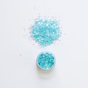 May include: A small jar of iridescent blue glitter with a small amount of glitter scattered around it on a white background.