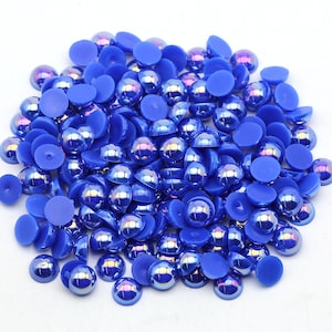 May include: A large pile of blue iridescent round plastic pearl cabochons. The cabochons are approximately 10mm in diameter and have a flat back.