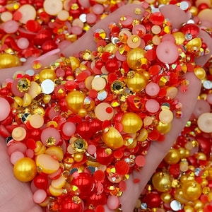 May include: A close-up of a variety of red and gold beads and rhinestones. The beads are arranged in a random pattern and are scattered across a hand.