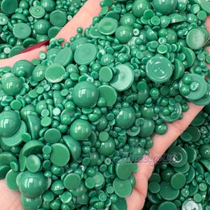 May include: A close-up shot of a handful of green wax beads in various shapes and sizes. The beads are a vibrant, glossy green color, with some appearing round and others flat. The image is well-lit, highlighting the texture and color of the wax.