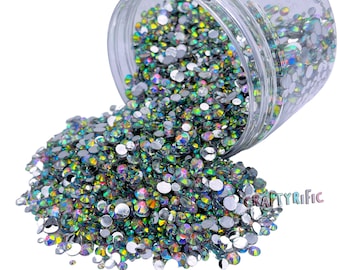 Pode incluir: Um frasco de plástico transparente inclinado, derramando uma grande quantidade de strass iridescentes. Os strass são redondos e refletem um arco-íris de cores. O frasco é parcialmente visível, e os strass estão espalhados sobre uma superfície branca. O texto "CRAFTYRIFIC" é visível.