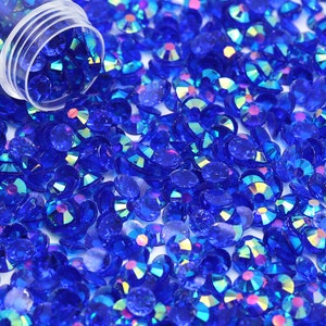 May include: A close-up image of a large quantity of small, iridescent blue rhinestones. The rhinestones are scattered on a white surface.