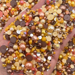 May include: A large assortment of brown, gold, and cream colored pearl and round resin embellishments. The embellishments are scattered on a hand.