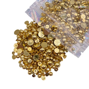 May include: A large quantity of gold-colored round and flat embellishments, scattered on a white surface. The embellishments are in a clear plastic bag.