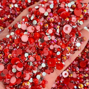 May include: A large assortment of red and pink round and flat rhinestones. The rhinestones are in various sizes and have a shiny, iridescent finish.