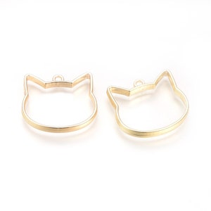 May include: Two gold metal cat head shaped pendant blanks with a loop at the top for hanging.