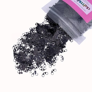 May include: A close-up of a bag of black glitter confetti. The confetti is shaped like handcuffs. The bag is labeled "CraftTribe" and has a pink background.