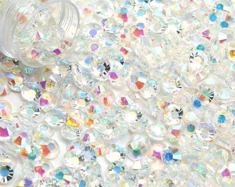 May include: A close-up image of a large quantity of iridescent clear glass rhinestones. The rhinestones are scattered on a white surface and are spilling out of a clear plastic container.