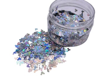 May include: A clear plastic jar filled with iridescent silver glitter shaped like Mickey Mouse heads. The glitter is spilling out of the jar onto a white background.